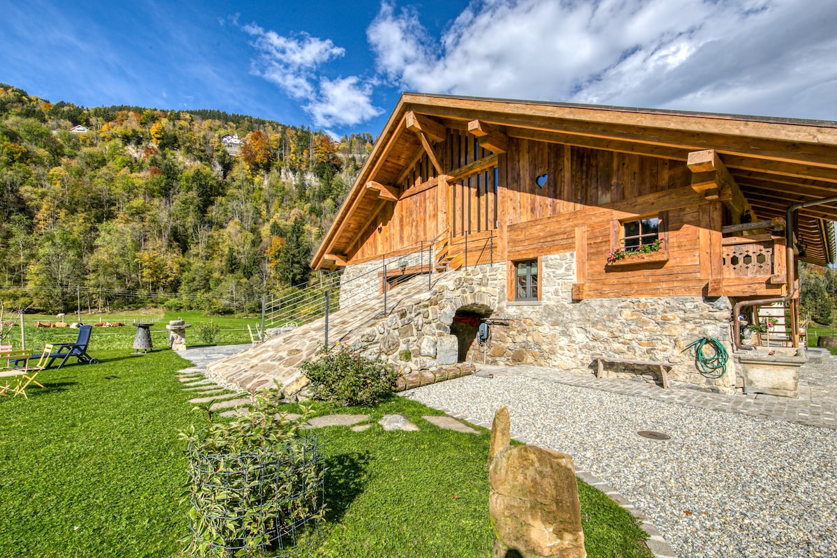 A wooden chalet with a welcoming stone foundation is set against a backdrop of trees displaying autumn colors. A landscaped area includes a grassy lawn and a gravel pathway leading to the entrance. Outdoor seating options create a relaxed outdoor atmosphere.