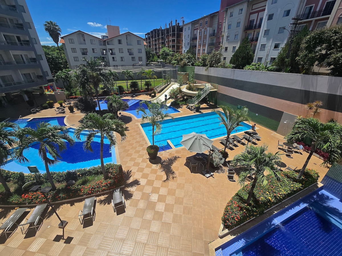 A spacious outdoor area is displayed, featuring multiple swimming pools, including three heated and three cold. Surrounding palm trees and well-maintained gardens enhance the environment. Lounge chairs and shaded umbrellas provide seating options around the pools, which are accompanied by a slide in the children's section.