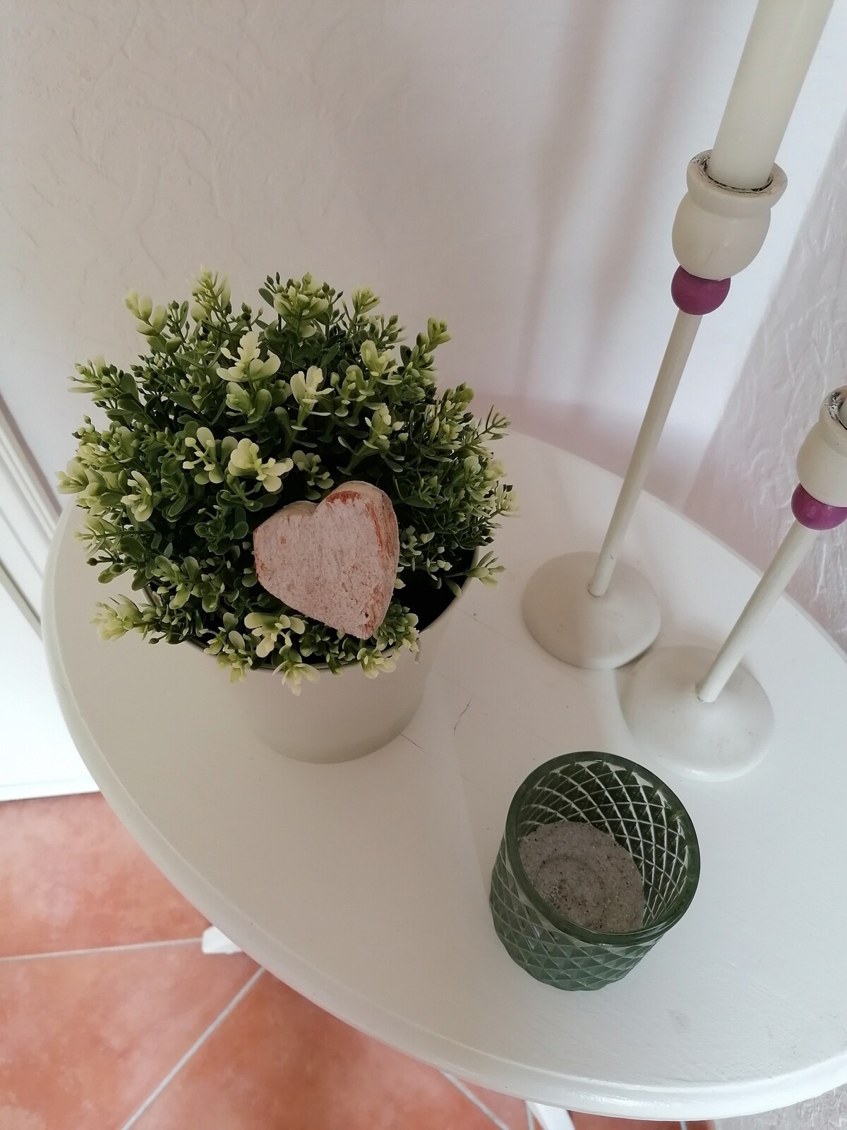 A small decorative table is featured, adorned with a potted plant that includes a heart-shaped tag. Two candle holders stand nearby, and a textured green glass item sits on the surface. The warm tile flooring complements the arrangement.