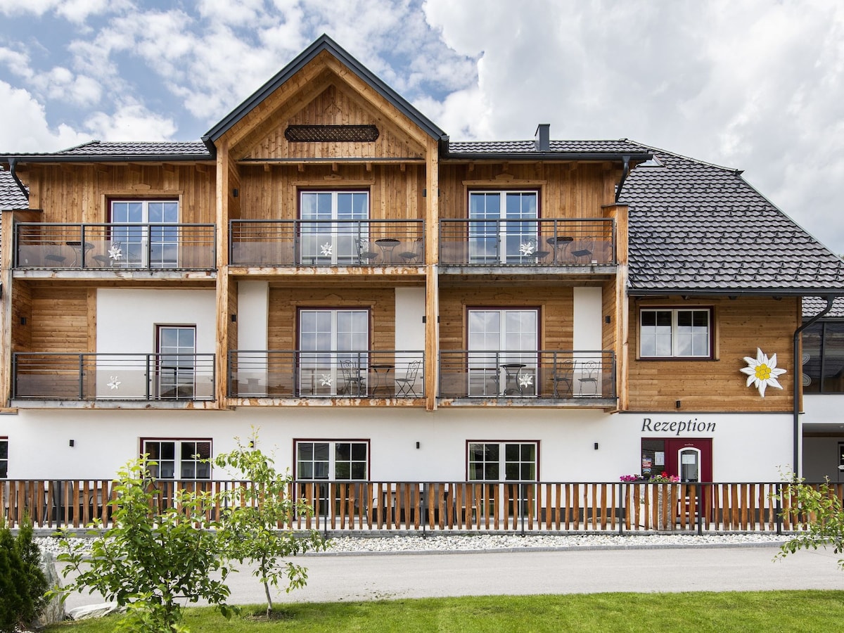 The exterior of a charming Alpine-style building features wooden balconies and intricate decorative elements. A reception area is indicated on one side, with a smooth driveway in front. Lush greenery is present, contributing to a welcoming and tranquil atmosphere.