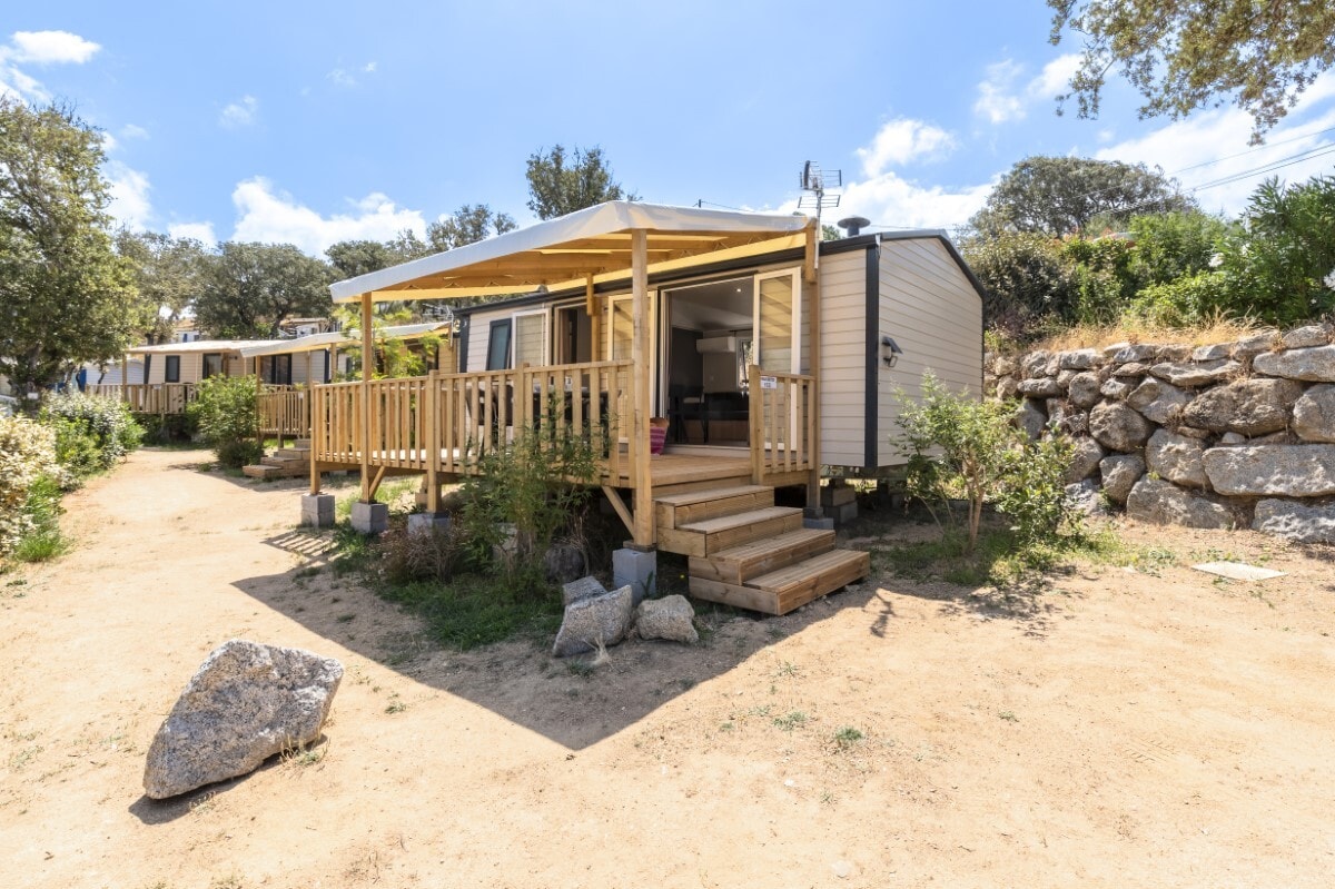 A mobile home is showcased with a wooden terrace, featuring steps leading to the entrance. The structure is surrounded by greenery and natural stone, set against a bright blue sky with scattered clouds.