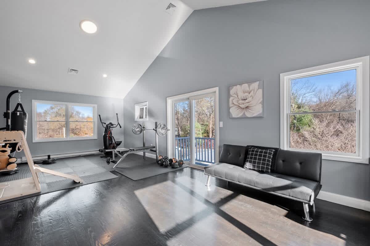 A bright bonus room features high ceilings and large windows, allowing natural light to fill the space. Gym equipment, including a stationary bike and weights, is neatly arranged alongside a cozy futon, creating an inviting area for both relaxation and activity.