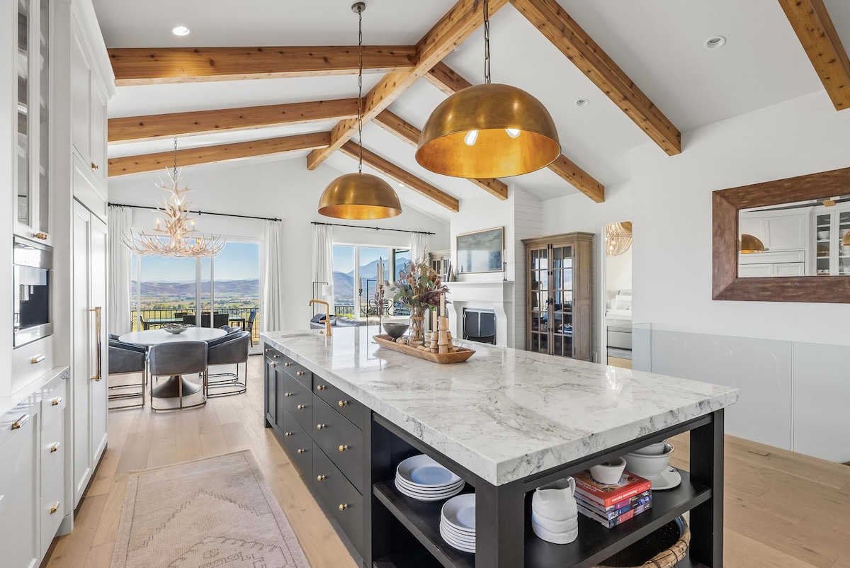 A spacious kitchen area showcases a large island topped with white marble, surrounded by dark cabinetry. Pendant lighting made of gold hangs above, illuminating the elegant design. Large windows invite natural light and offer scenic views of the surrounding landscape, enhancing the open-concept layout.