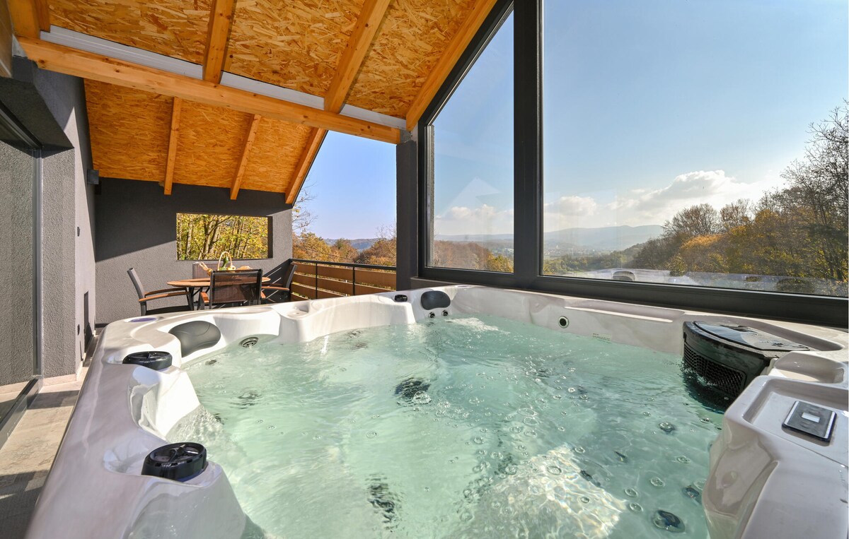 A heated outdoor hot tub is set under a wooden ceiling, featuring multiple jets and a spacious design. Large windows offer expansive views of the surrounding hills and valleys, allowing natural light to fill the area. A dining table and chairs are visible nearby.