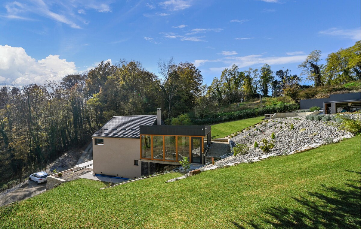 A modern holiday home is situated on a hillside, featuring a combination of sleek design and natural surroundings. Large windows provide an open view of the landscape, while the extensive green lawn leads to terraced areas adorned with rocks and plants.