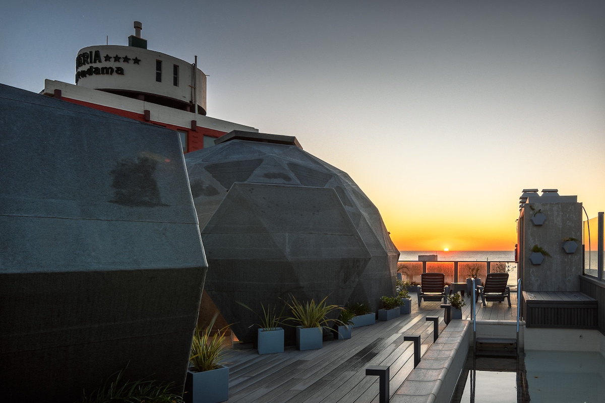 A modern terrace features unique geometric domes overlooking a sunset view. Lounge chairs are positioned along a wooden walkway, with potted plants enhancing the serene outdoor space. The distant ocean reflects warm hues, creating a tranquil ambiance for relaxation.