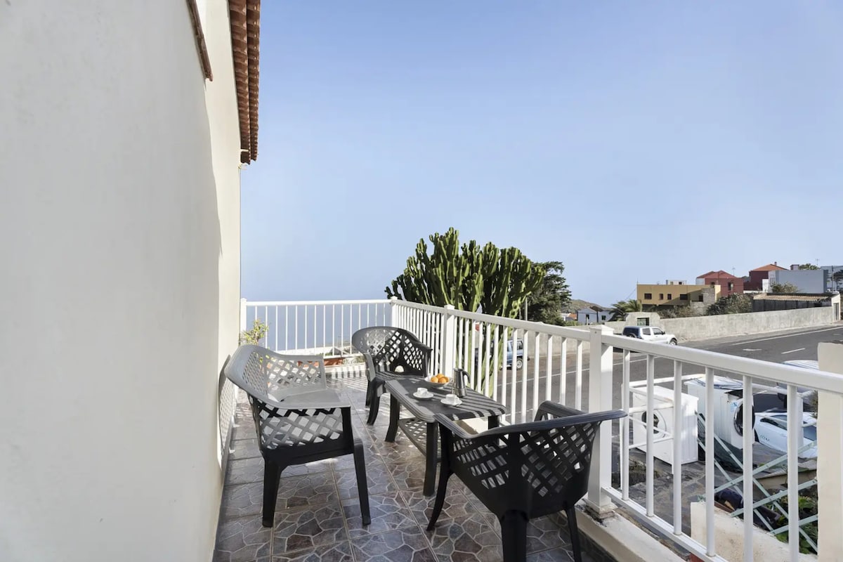 A private terrace is featured, furnished with four black chairs and a table. The view includes a distant coastline and surrounding greenery. The floor showcases patterned tiles, providing a comfortable outdoor space for relaxation.