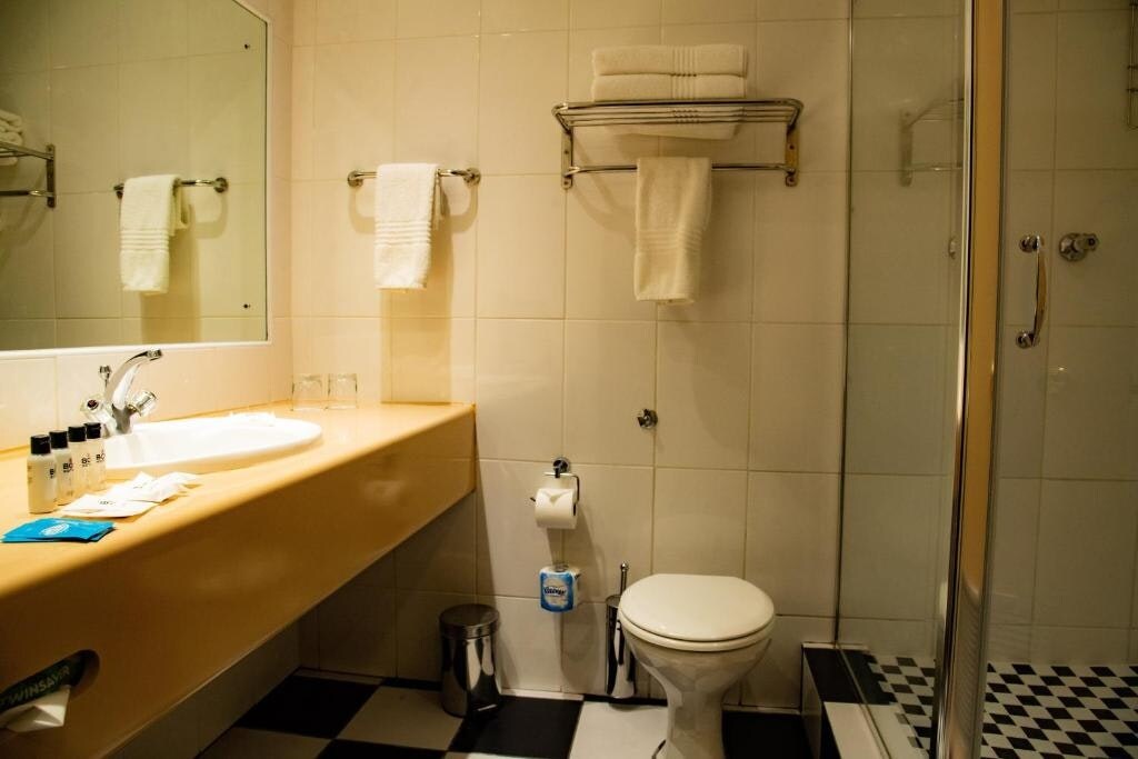 The bathroom features a modern design with a wall-mounted sink and a large mirror above. Shelving holds neatly arranged towels, while personal care products are displayed nearby. A walk-in shower is visible, alongside a toilet and a stylish black and white checkered floor.