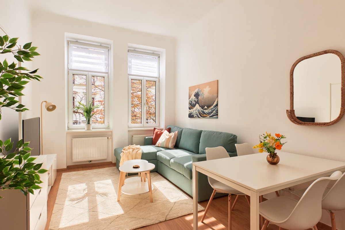 A bright living area features a soft green sofa accompanied by decorative cushions, a small white coffee table, and a dining table with four chairs. Large windows allow natural light to fill the space, complemented by a wall art piece depicting waves.