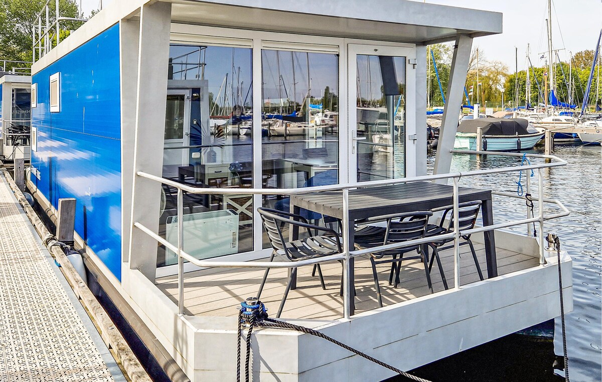 The houseboat features a spacious outdoor terrace with a dining table and chairs, offering views of the marina. Sunlight brightens the modern, blue exterior, while the surrounding boats add to the tranquil harbor atmosphere.