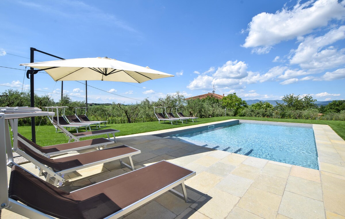 A private outdoor pool is surrounded by a well-manicured lawn and olive trees. Comfortable lounge chairs are set by the poolside, with a large umbrella providing shade. Expansive skies and a few scattered clouds create a serene atmosphere.