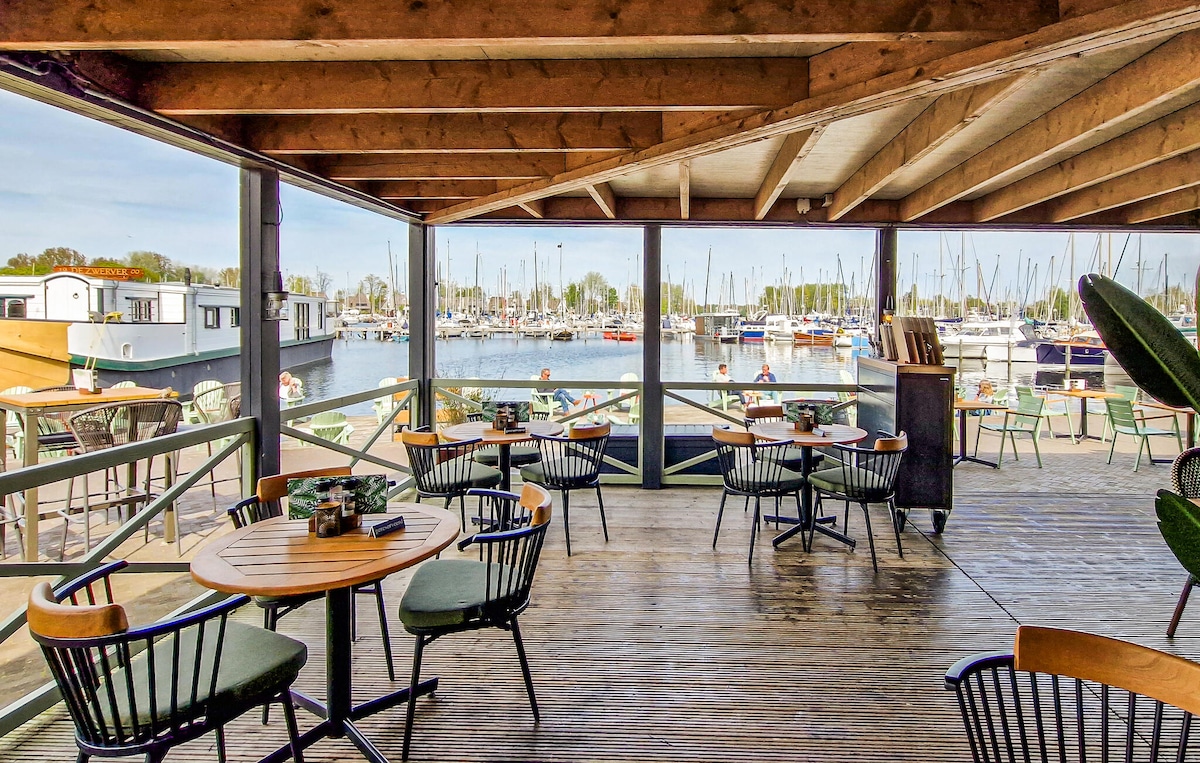 An open terrace features several round tables and chairs with views overlooking the harbour filled with boats. Large windows provide natural light, and the wooden beams in the ceiling enhance the spacious feel of the area.