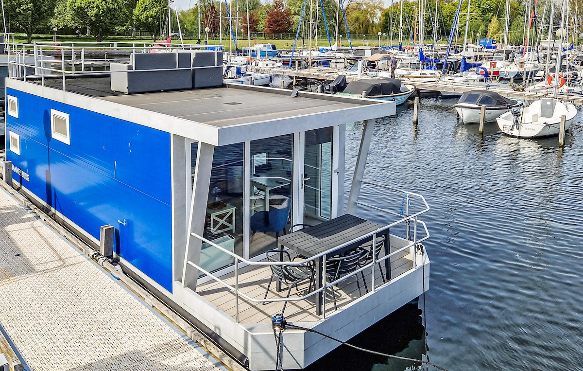 The houseboat features a modern exterior with a vibrant blue facade and a spacious terrace overlooking the harbour. Outdoor seating is available, allowing for relaxed meals or gatherings while enjoying views of the calm waters and surrounding marina.