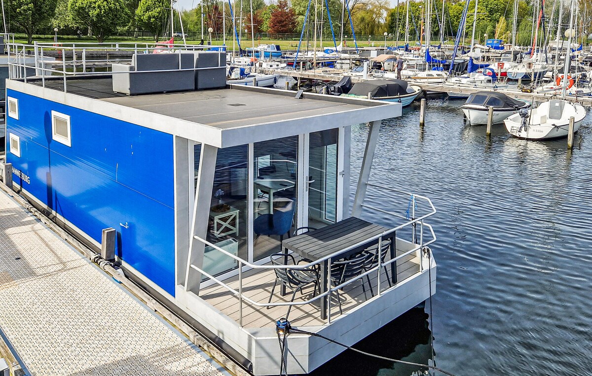 A bright blue houseboat sits moored in the harbor, featuring an outdoor terrace with a large table and chairs. The surrounding water reflects the sky, while various boats are docked nearby. Lush greenery lines the shore, offering a calm and tranquil setting.