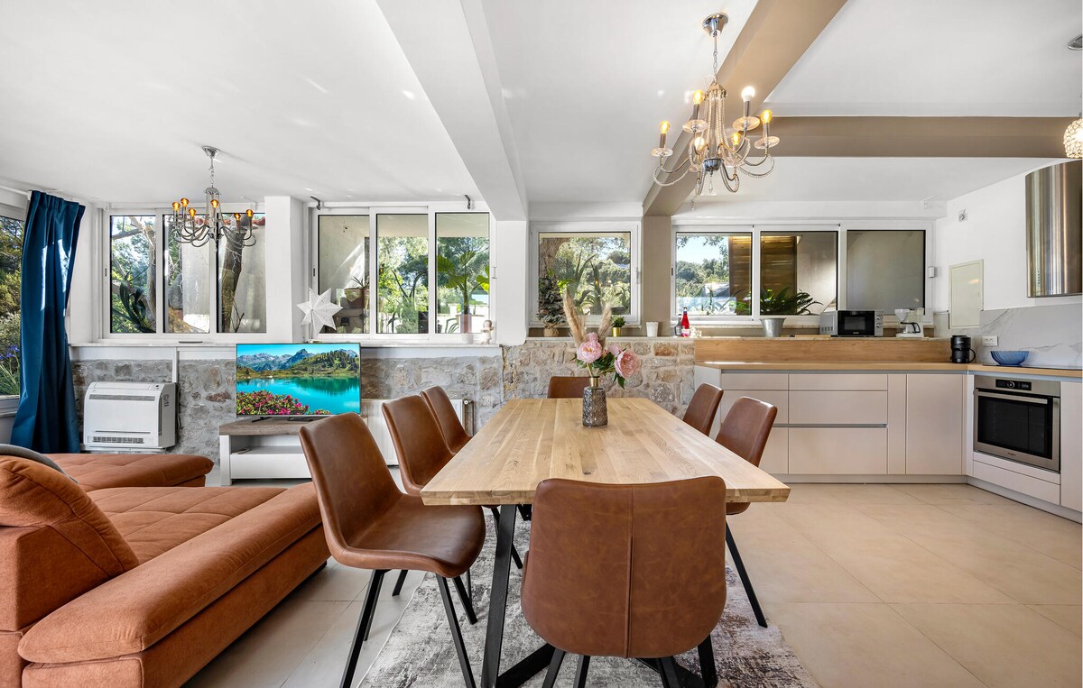 A spacious dining area features a wooden table surrounded by brown chairs. Large windows allow natural light to fill the room, and a cozy sofa is positioned nearby. A modern kitchen with sleek appliances and a vibrant artwork adds warmth to the serene space.