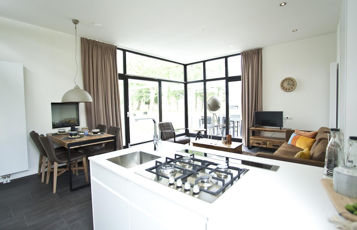 The open kitchen features a modern gas stove and sleek white cabinetry. A spacious dining table with several chairs sits adjacent, while a cozy seating area with a television is positioned nearby. Large windows allow natural light to illuminate the space and offer views of the outside.
