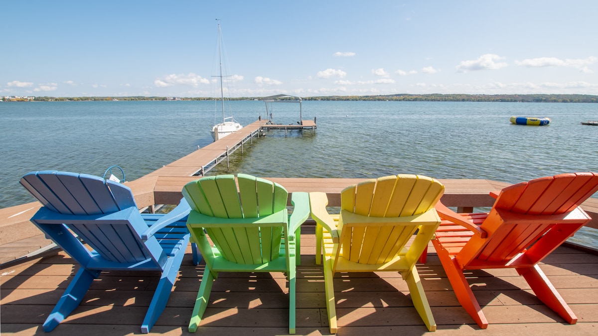 A set of four colorful adirondack chairs in blue, green, yellow, and orange is positioned on a wooden deck overlooking a serene lake. A dock stretches into the water, with a sailboat moored nearby, and distant hills can be seen against a clear blue sky.