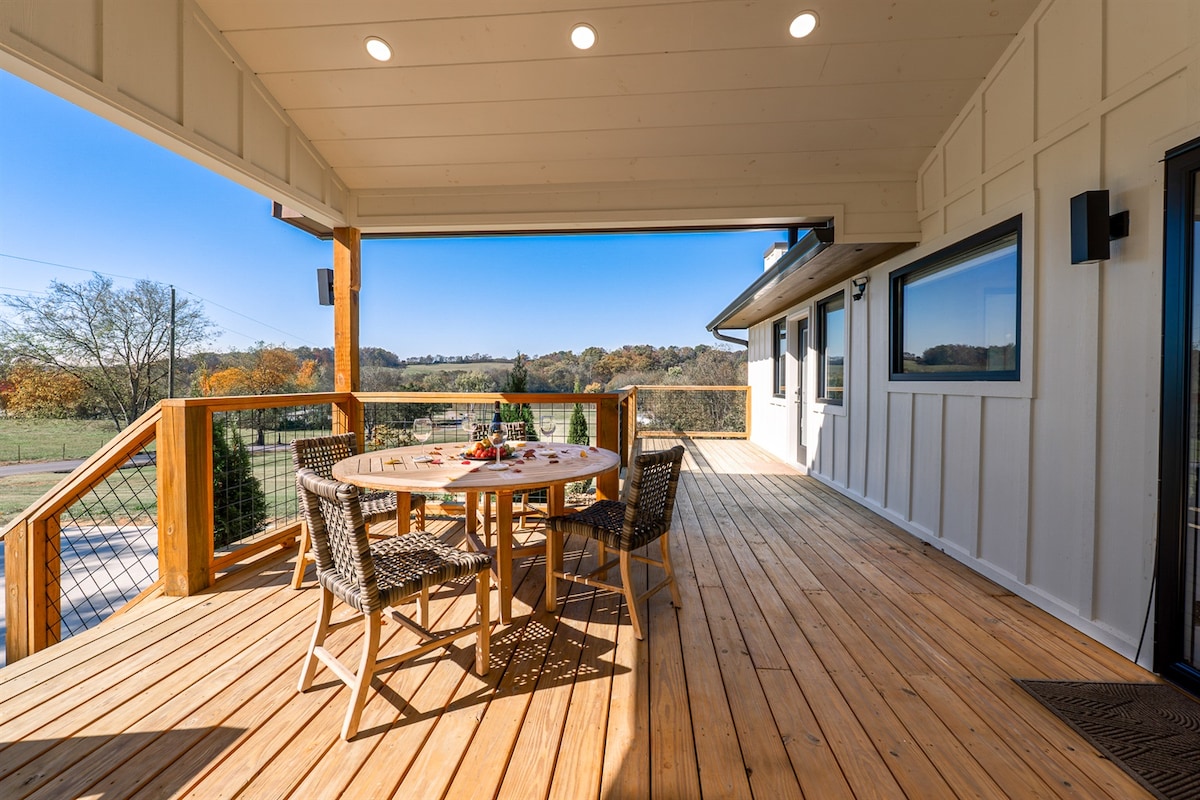 An outdoor deck area features a round wooden dining table surrounded by four woven chairs. Natural wood flooring enhances the inviting atmosphere, while expansive views of the rolling countryside and clear blue sky can be seen in the background.