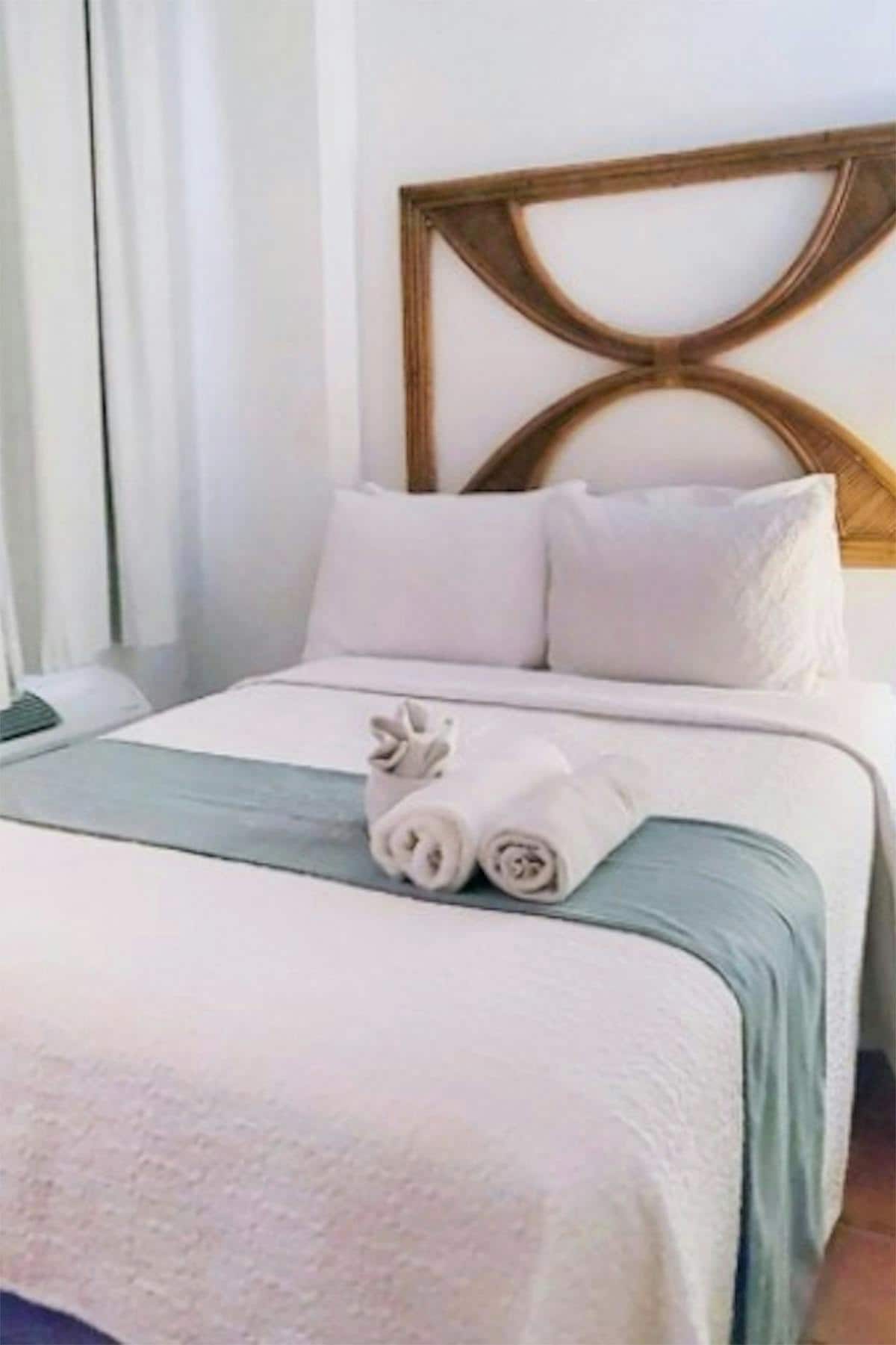 A cozy sleeping area is presented with a queen-sized bed, featuring a decorative headboard and crisp linens. A soft blanket drapes gently over the bed, accompanied by neatly rolled towels. Natural light filters through the window, highlighting the warm wooden floors.