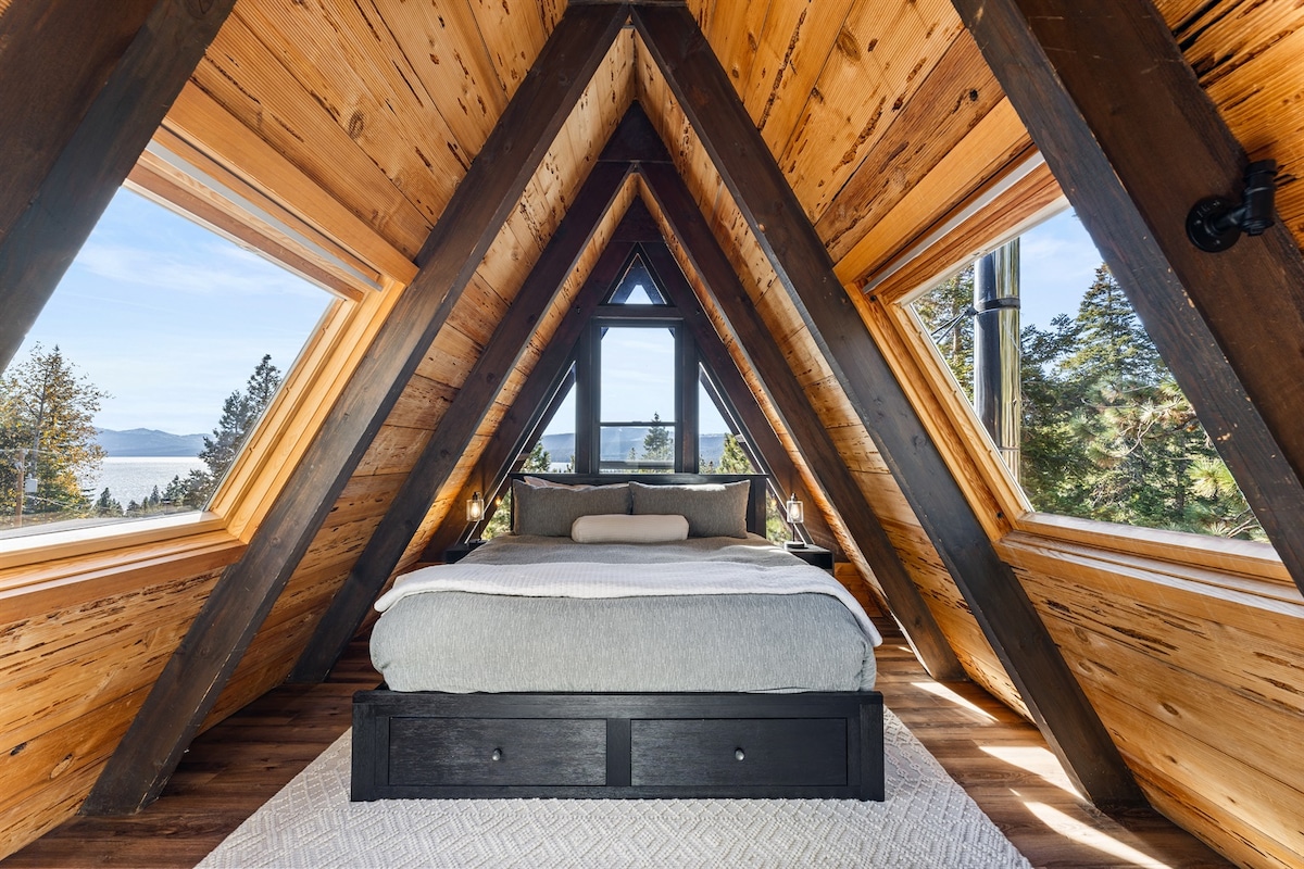 An inviting bedroom features a spacious bed framed by large triangular windows, offering picturesque views of Lake Tahoe. Natural wood paneling complements the cozy ambiance, creating a serene sleeping environment. The room is designed to maximize natural light and scenic outdoor vistas.