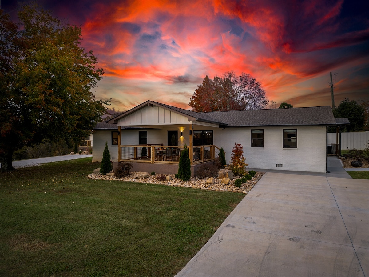 The home's exterior is showcased under a vibrant sunset sky, highlighting the well-maintained lawn and thoughtfully landscaped garden. A spacious wooden deck is visible, surrounded by small shrubs, and the paved driveway offers ample parking space next to the welcoming entrance.