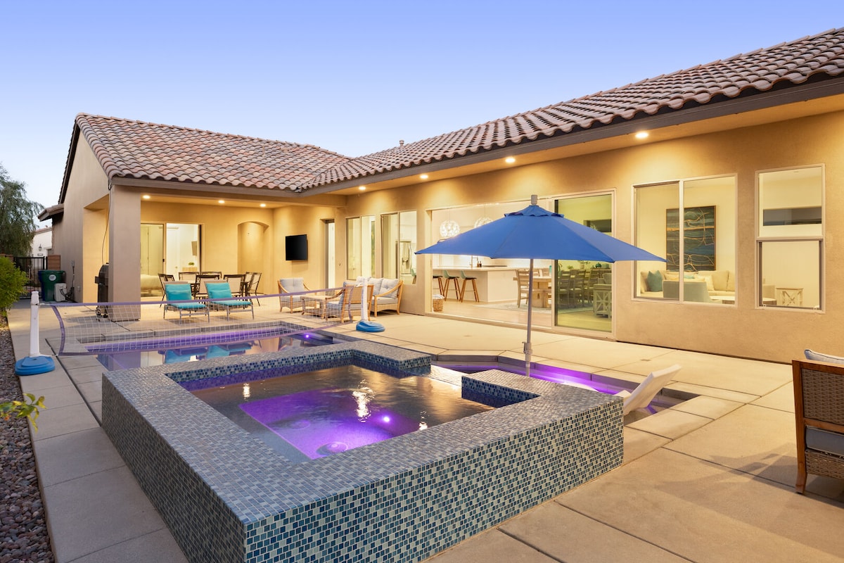 The private backyard features a hot tub and saltwater pool with radiant purple lighting. Poolside loungers are positioned under a large umbrella, while an outdoor dining area is visible through expansive glass sliding doors. The modern architecture complements the tranquil atmosphere.