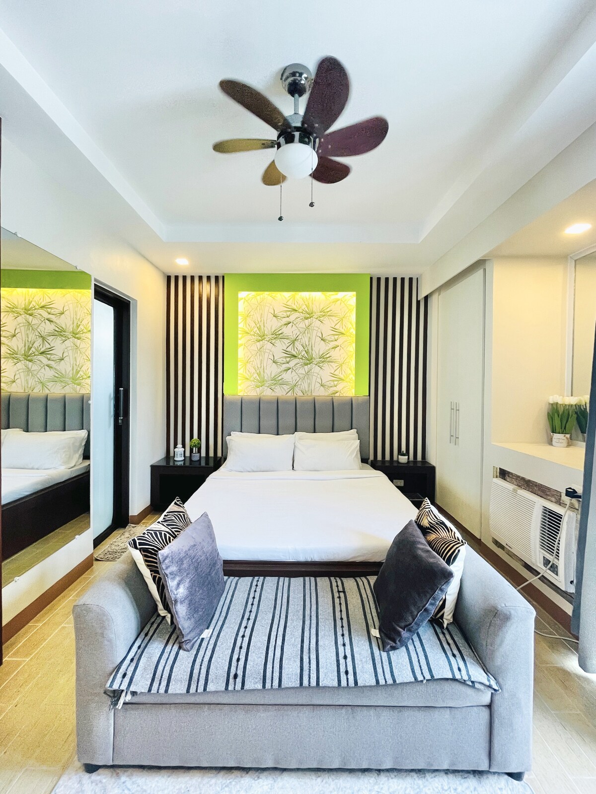 A modern studio interior features a spacious bed, complemented by decorative pillows on a sleek sofa. A vibrant wall art piece with green tones adds a touch of color. The room is brightened by natural light from the window, and light wood finishes enhance the overall ambiance.