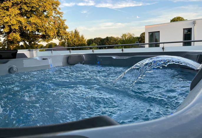 A hot tub is filled with clear, bubbling water, set against a backdrop of trees and a bright sky. Sunlight reflects on the water's surface, creating a sense of relaxation in this inviting outdoor space.