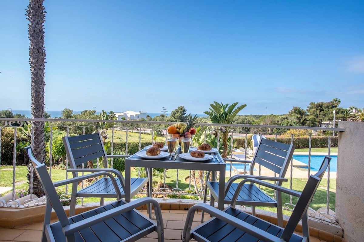 A private balcony is shown with two chairs and a small table, set with fruit and pastries. The view includes landscaped gardens and a swimming pool, with the ocean visible in the distance under a clear blue sky.