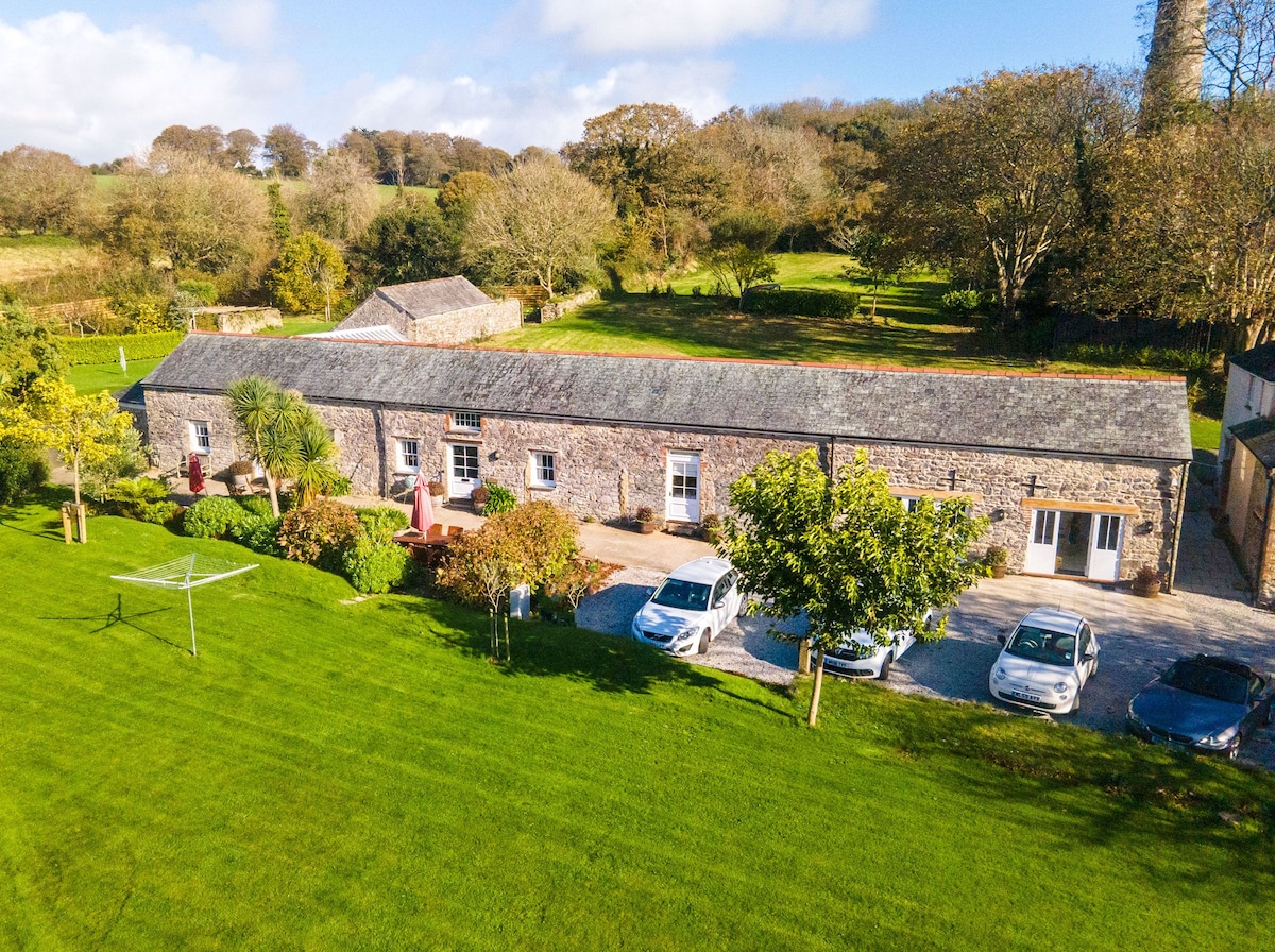 The exterior of Dolphin Cottage showcases a stone barn structure surrounded by lush green lawns. Several vehicles are parked in front. A walkway leads to the entrance, while trees and shrubs enhance the landscape, creating a welcoming ambiance within the Cornish countryside.