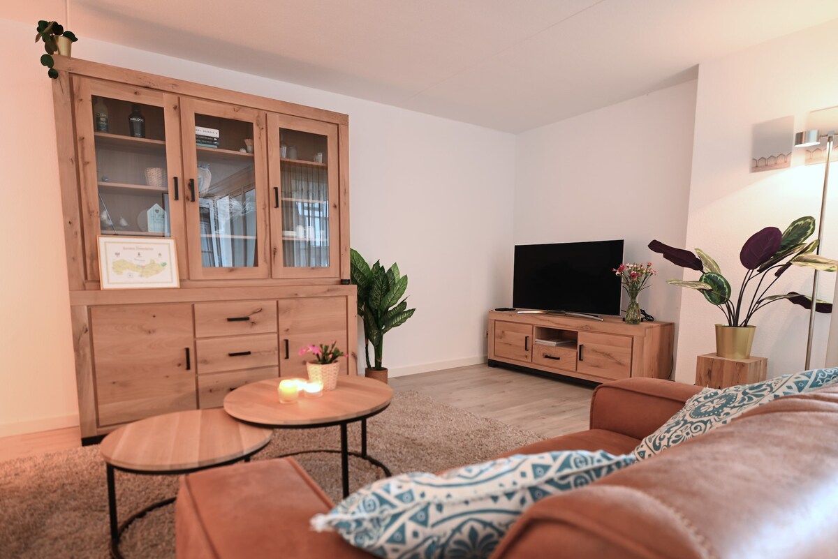 A cozy living area is presented with a wooden cabinet featuring glass doors, displaying decorative items. A flat-screen television is positioned on a wooden media stand. Two round coffee tables sit on a plush area rug, accompanied by a green plant and a vase of flowers.