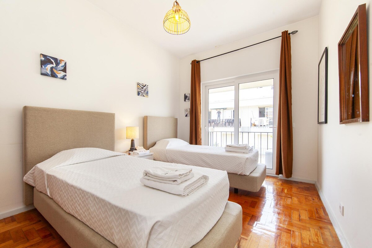 The private bedroom features two single beds with light bedding, positioned on a wooden floor. A bedside lamp is present, and artwork decorates the walls. Natural light fills the space through a large glass door that opens to the small private balcony.