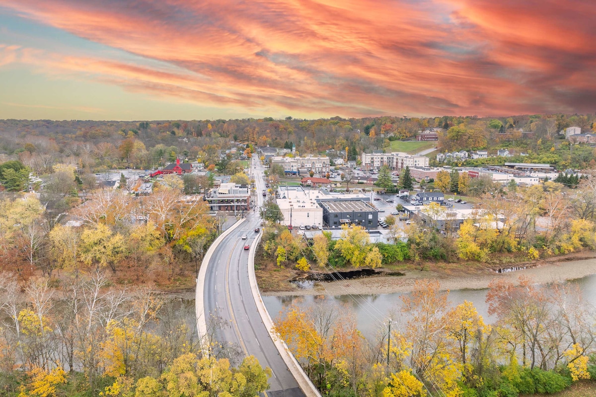 An aerial view captures the scenic town of Loveland, Ohio, highlighting a winding road that crosses over a river. Fall foliage paints the trees in vibrant hues, while warm colors fill the sky during sunset, creating a peaceful atmosphere over the charming community and surrounding nature.