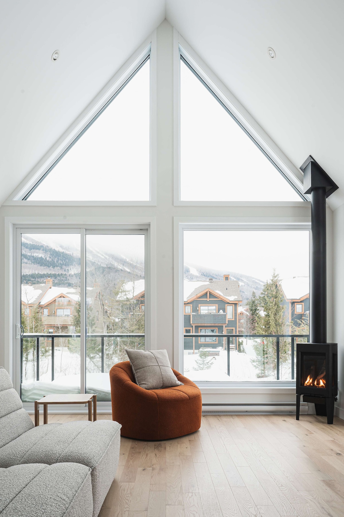 A bright living area features tall triangular windows allowing natural light and scenic views of the snowy landscape. A cozy orange armchair and a light grey sectional sofa are arranged near a modern gas fireplace, creating a comfortable seating arrangement.