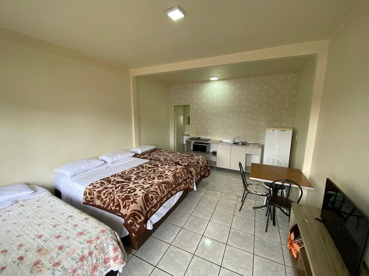 The spacious loft features multiple beds covered with patterned bedding. A small dining table with chairs is positioned near a kitchenette equipped with essential appliances, including a stove, microwave, and refrigerator. A television is placed on a low stand against a wall.