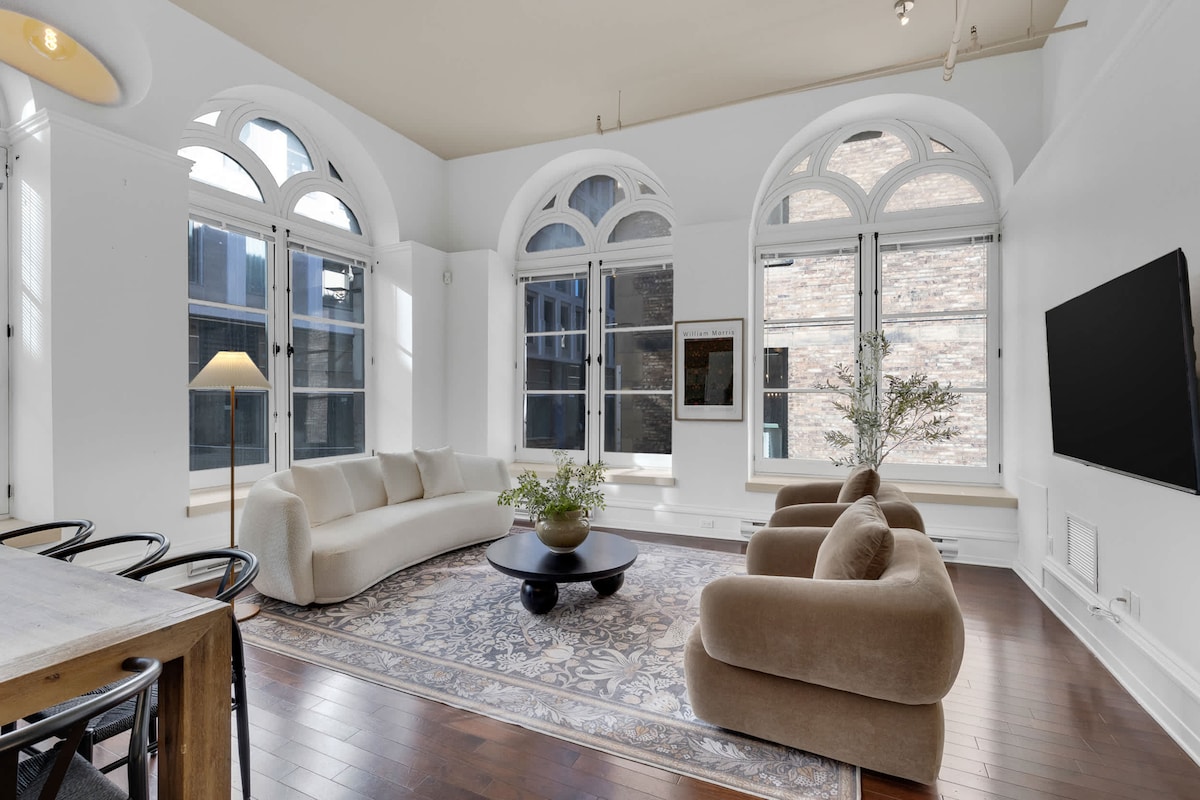 A spacious living area features large arched windows that allow natural light to fill the room. A light-colored, curved sofa is paired with two plush armchairs, centered around a round coffee table. A large TV is mounted on the wall, with a delicate rug anchoring the space.