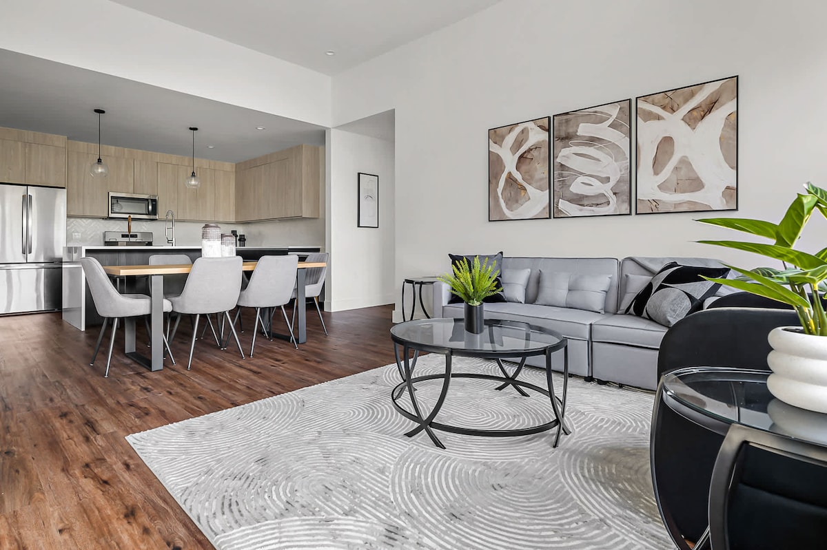 The open living area features a comfortable sofa positioned near a circular coffee table. A modern dining table with several chairs is visible in the background. Light-colored cabinetry and stainless steel appliances enhance the kitchen space. Artistic wall decor adds a touch of character.