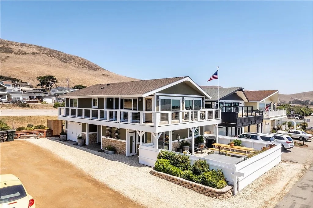 The exterior of The Cody House features a two-story structure set against a coastal backdrop, with a welcoming porch and American flag flying. A spacious gravel area accommodates parking, while nearby, an outdoor dining area with a large picnic table is visible, offering a perfect spot for gatherings.