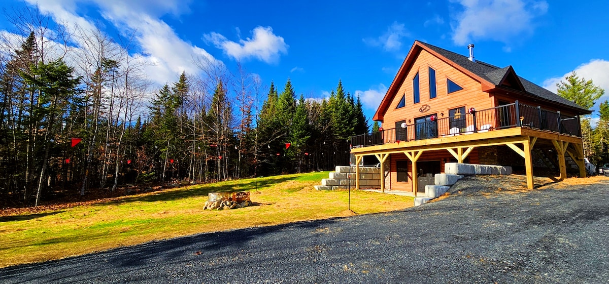 The cabin, constructed from warm-toned wood, features a welcoming front porch supported by sturdy pillars. A large deck extends from the main entrance, offering a view of the surrounding trees and grassy area. An outdoor fire pit, surrounded by seating, is visible on the lawn.