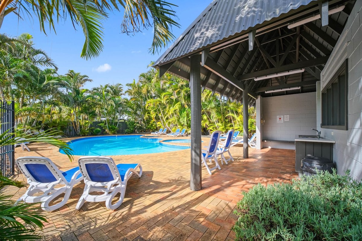 A refreshing pool area is framed by lush palm trees, creating a serene backdrop. Sun loungers are arranged nearby, offering relaxation space. An outdoor kitchen is positioned under a shaded pavilion, providing convenience for gatherings. The blue water of the pool invites leisure and enjoyment.