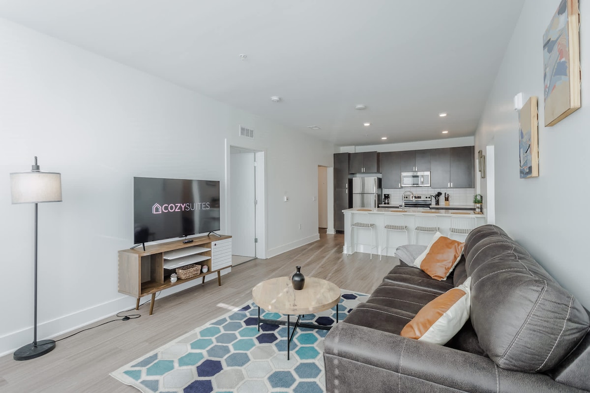 A spacious living area features a modern sofa with colorful cushions, a large area rug with geometric patterns, and a coffee table placed centrally. A flat-screen TV is mounted on the wall opposite the seating. The kitchen area is visible, showcasing sleek cabinets and stainless-steel appliances.