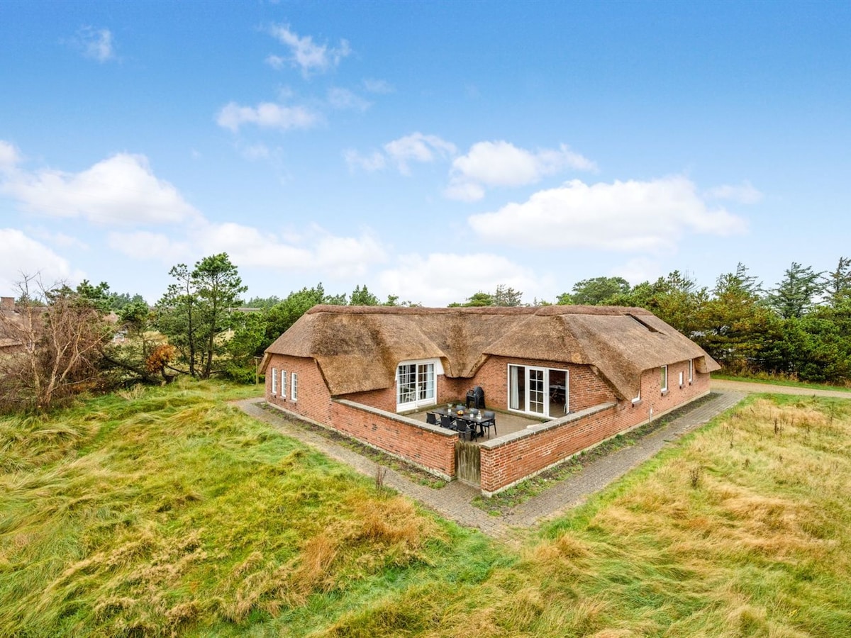 The exterior of a spacious five-room house is shown, featuring a traditional thatched roof and brick facade. Surrounding greenery and a natural landscape complement the inviting setting. A pathway leads to a patio area where outdoor seating is visible.