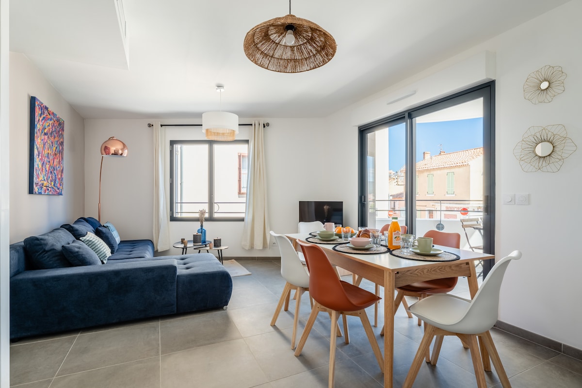 A bright living area features a comfortable blue sofa and a modern dining table set for four. Large sliding glass doors provide outdoor access and natural light, with framed artwork adding a pop of color to the neutral walls. A flat-screen TV is positioned near the dining setup.
