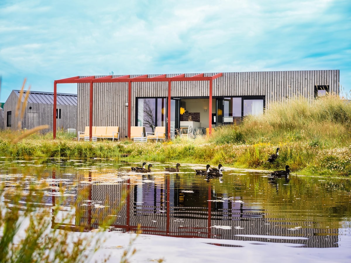 The modern lodge features a sleek wooden exterior and red accents, surrounded by lush greenery and a tranquil pond. The reflection of the lodge is visible in the water, while ducks can be seen leisurely swimming, creating a serene outdoor scene.
