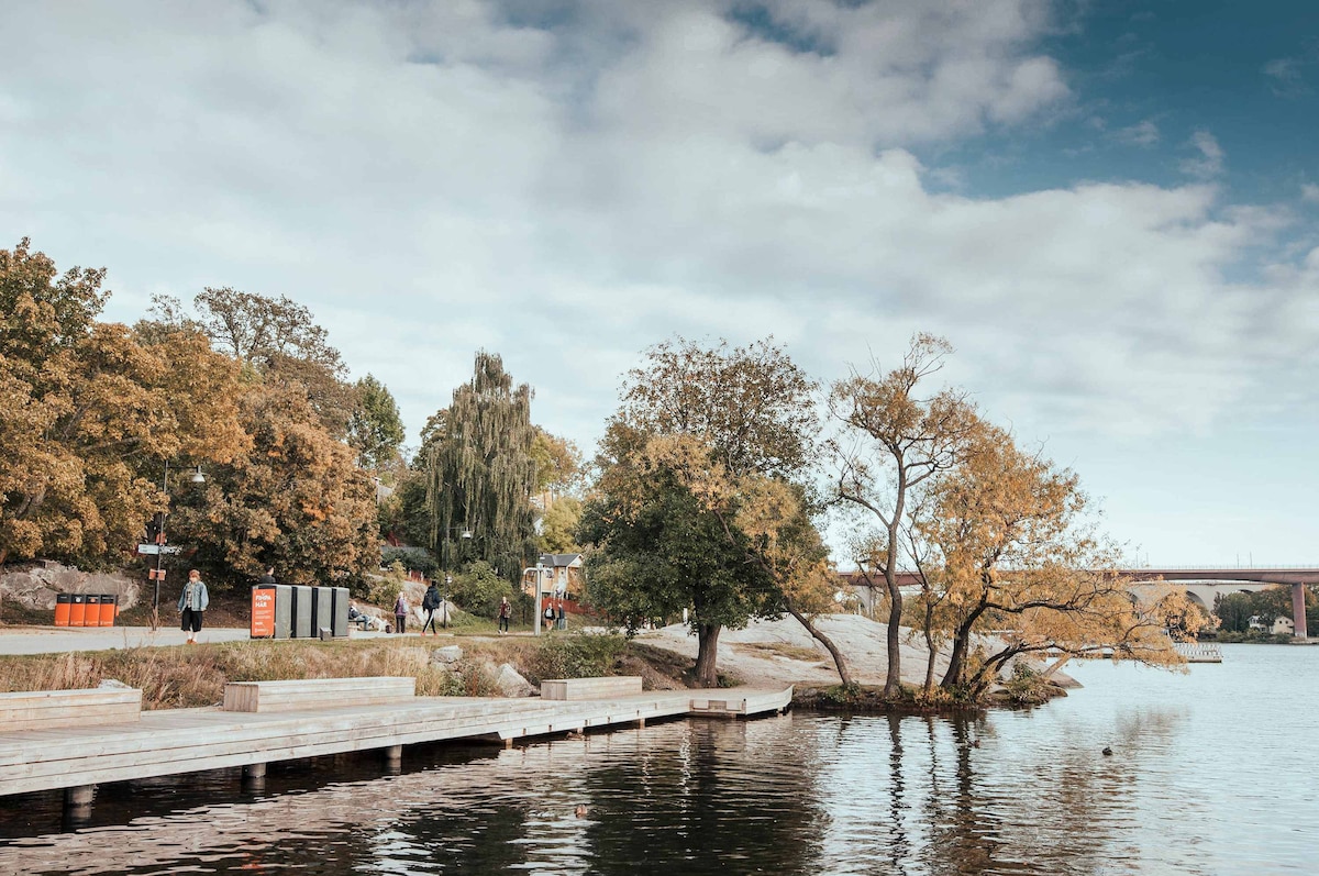 A serene waterfront view captures trees with autumn foliage along the banks. A wooden dock extends into the water, where gentle ripples reflect the sky. People are seen strolling in the nearby park, maintaining a peaceful atmosphere.