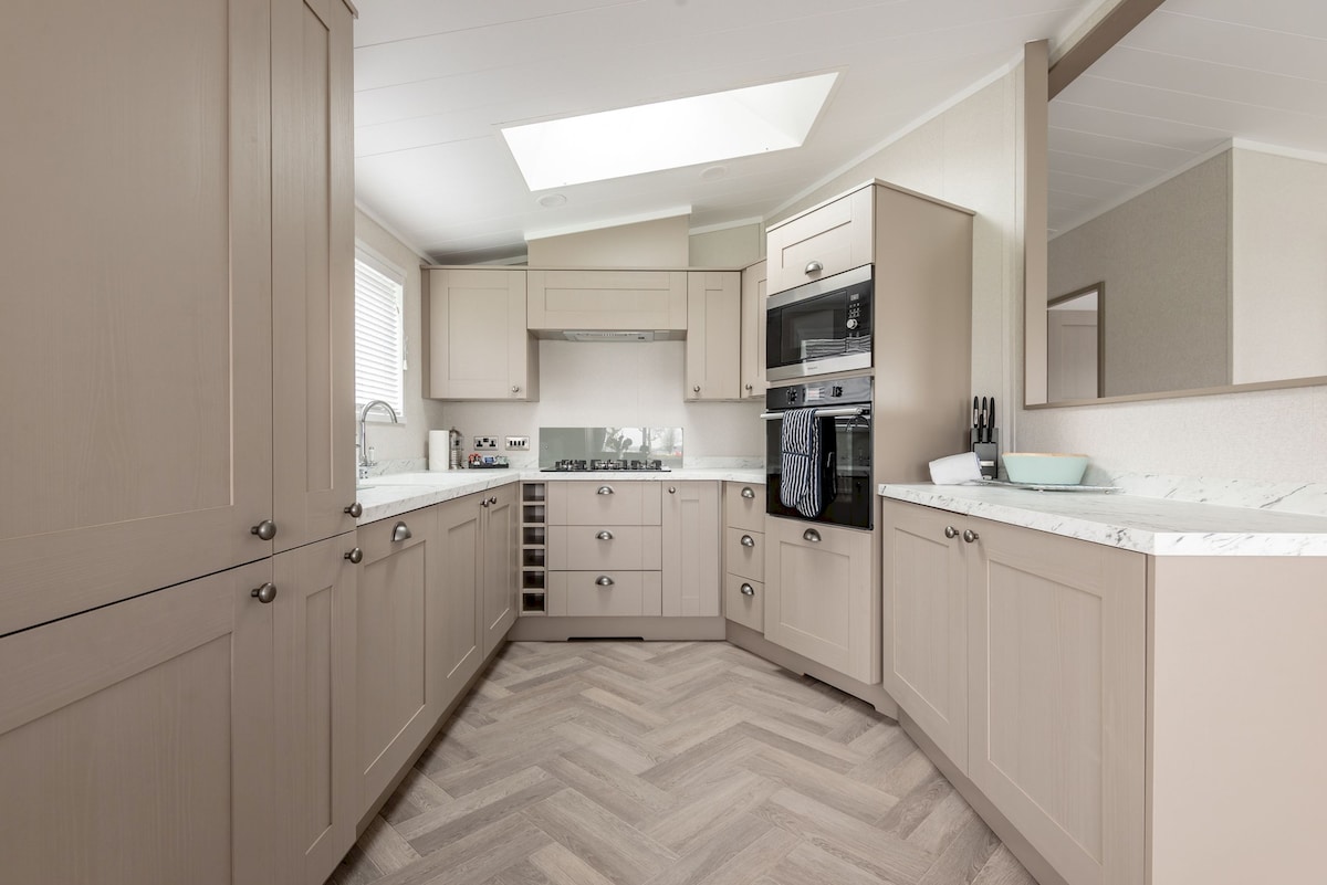 A modern kitchen showcases light-colored cabinetry and a marble countertop. Appliances including an oven, microwave, and wine cooler are neatly integrated into the design. Natural light floods the space through a skylight, highlighting the herringbone-patterned flooring.