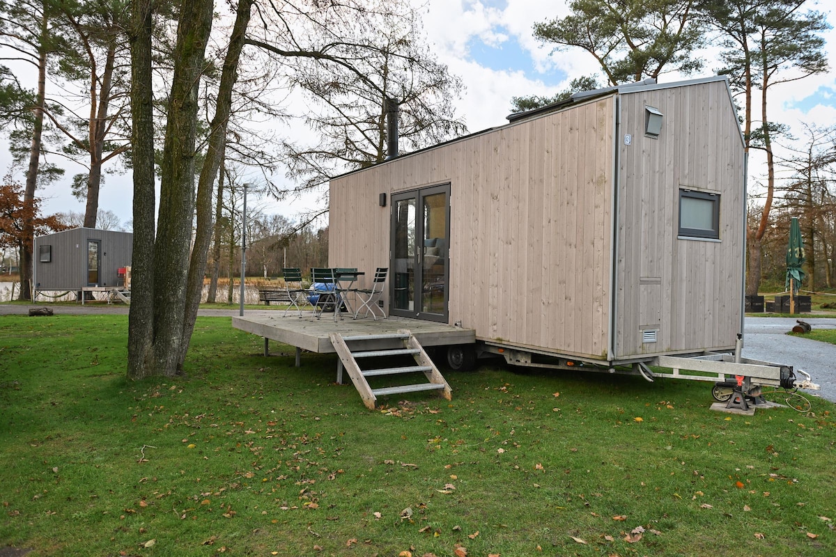 A compact wooden house is positioned on a grassy area, featuring a small deck with outdoor seating arrangements. Large windows allow for natural light, while nearby trees provide a sense of privacy. The surrounding landscape includes additional tiny houses in the background.