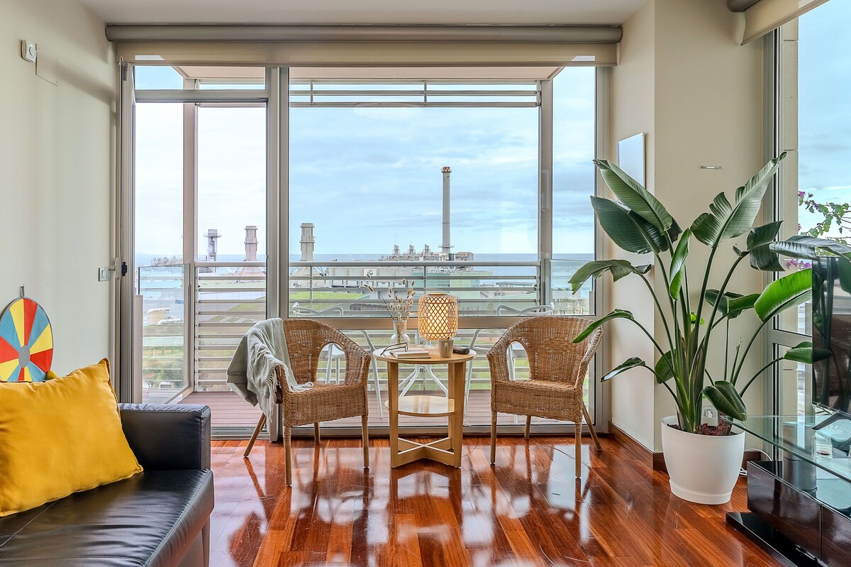 A cozy living area features two woven chairs and a small table on a wooden deck, surrounded by large windows offering a view of the outdoors. A vibrant green plant adds a natural touch to the space, complemented by warm wood flooring.
