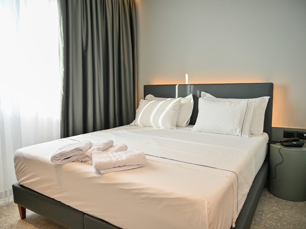 A well-appointed sleeping area featuring a spacious bed adorned with crisp white linens and plush pillows. Soft natural light filters through sheer curtains, while a bedside table holds neatly arranged towels. Elegant gray curtains frame the window, enhancing the room's modern design.