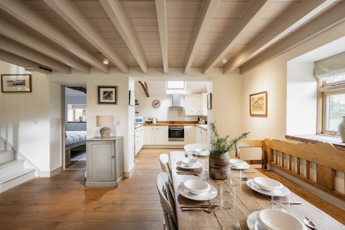 The spacious kitchen and dining area is highlighted by rustic wooden beams above. A large oak dining table is set with plates, inviting shared meals. The kitchen features modern appliances and a warm, welcoming layout, ensuring a comfortable space for cooking and dining.